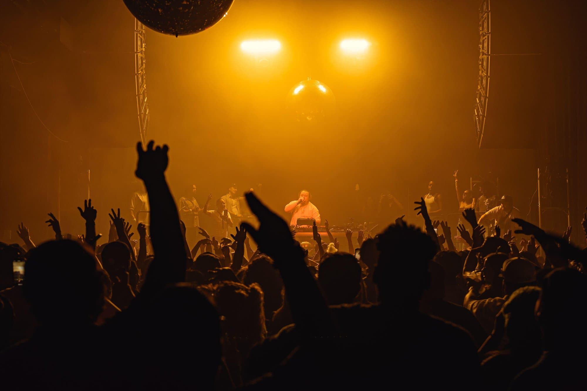 Hands in the air facing a DJ under a mirror ball in amber light