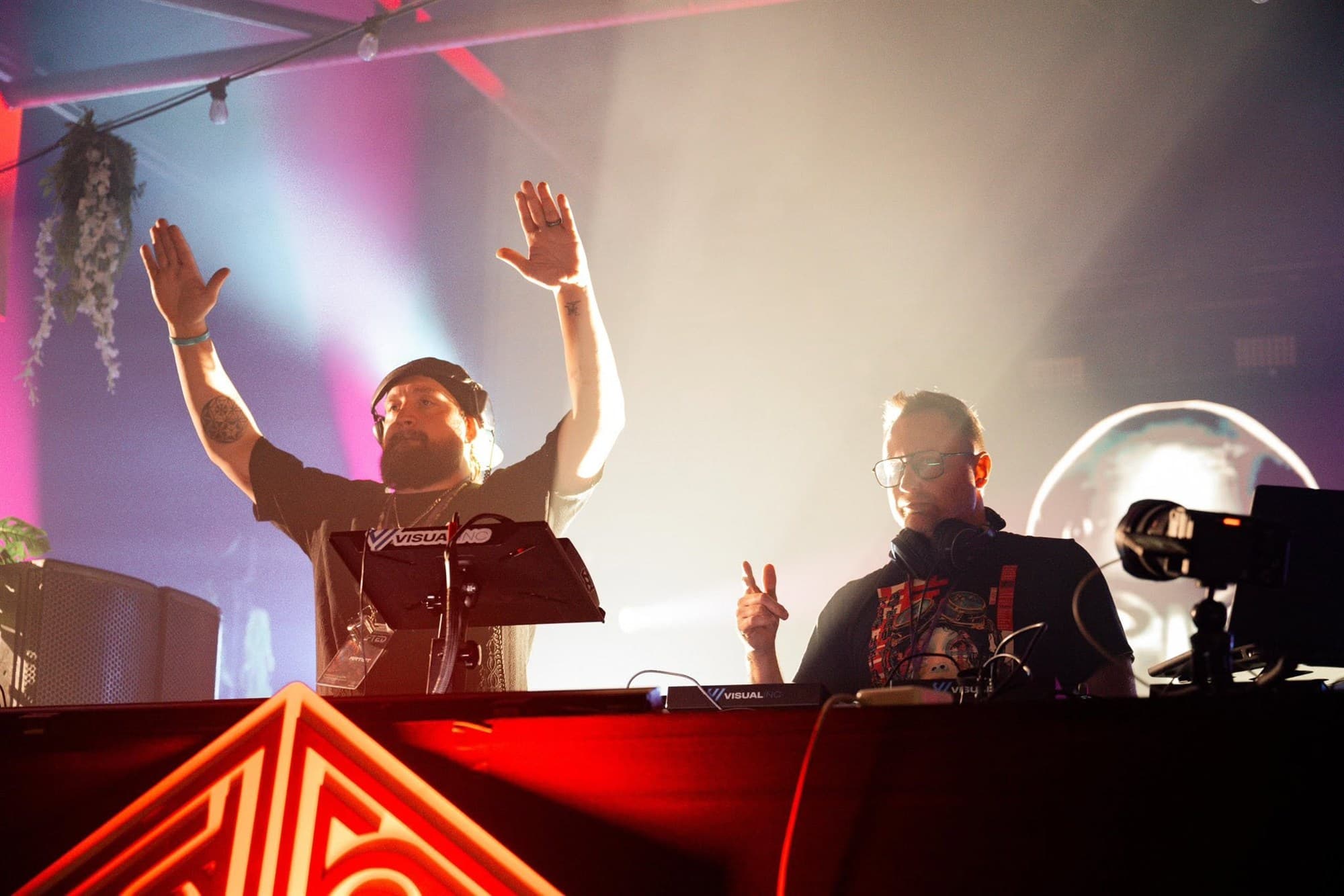 Two DJs behind the decks with their hands in the air under pink and blue stage lights and fog