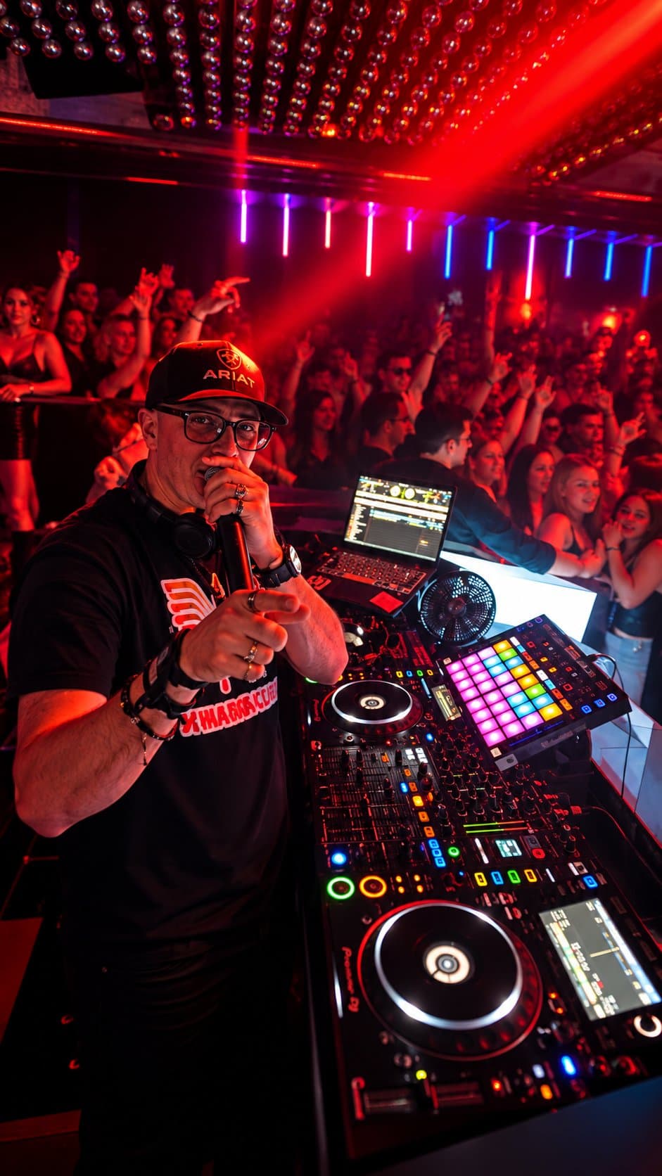 DJ with a microphone behind a Pioneer rig with a crowd in red light